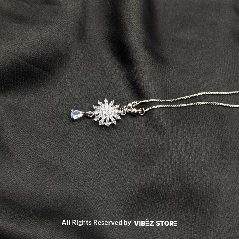 Shimmering Starburst Locket with Teardrop Pendant 2 A close-up of a silver-tone starburst locket adorned with shimmering crystals and a teardrop-shaped gemstone, laid on a black fabric background.