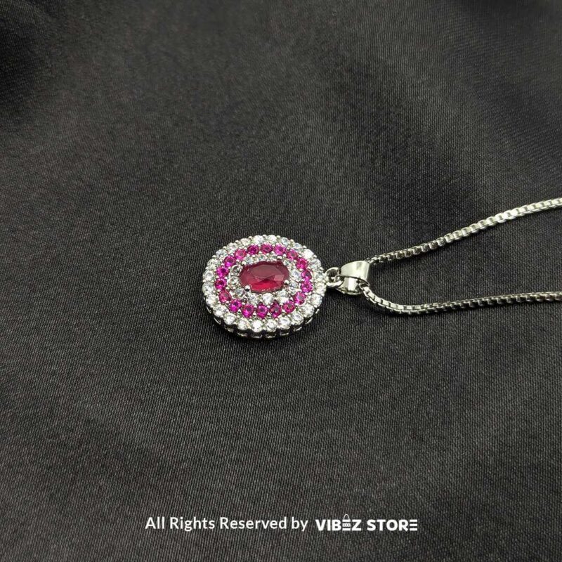 Pink Gemstone Halo Locket 1 A silver-tone locket featuring a pink gemstone surrounded by a double halo of crystals, displayed on a black fabric background.